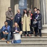 group of eight delegates pose on the stone steps beneath the columned entrance of the British Museum, some standing and some seated with bags.