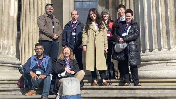 group of eight delegates pose on the stone steps beneath the columned entrance of the British Museum, some standing and some seated with bags.