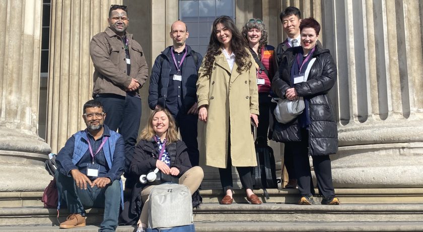 group of eight delegates pose on the stone steps beneath the columned entrance of the British Museum, some standing and some seated with bags.